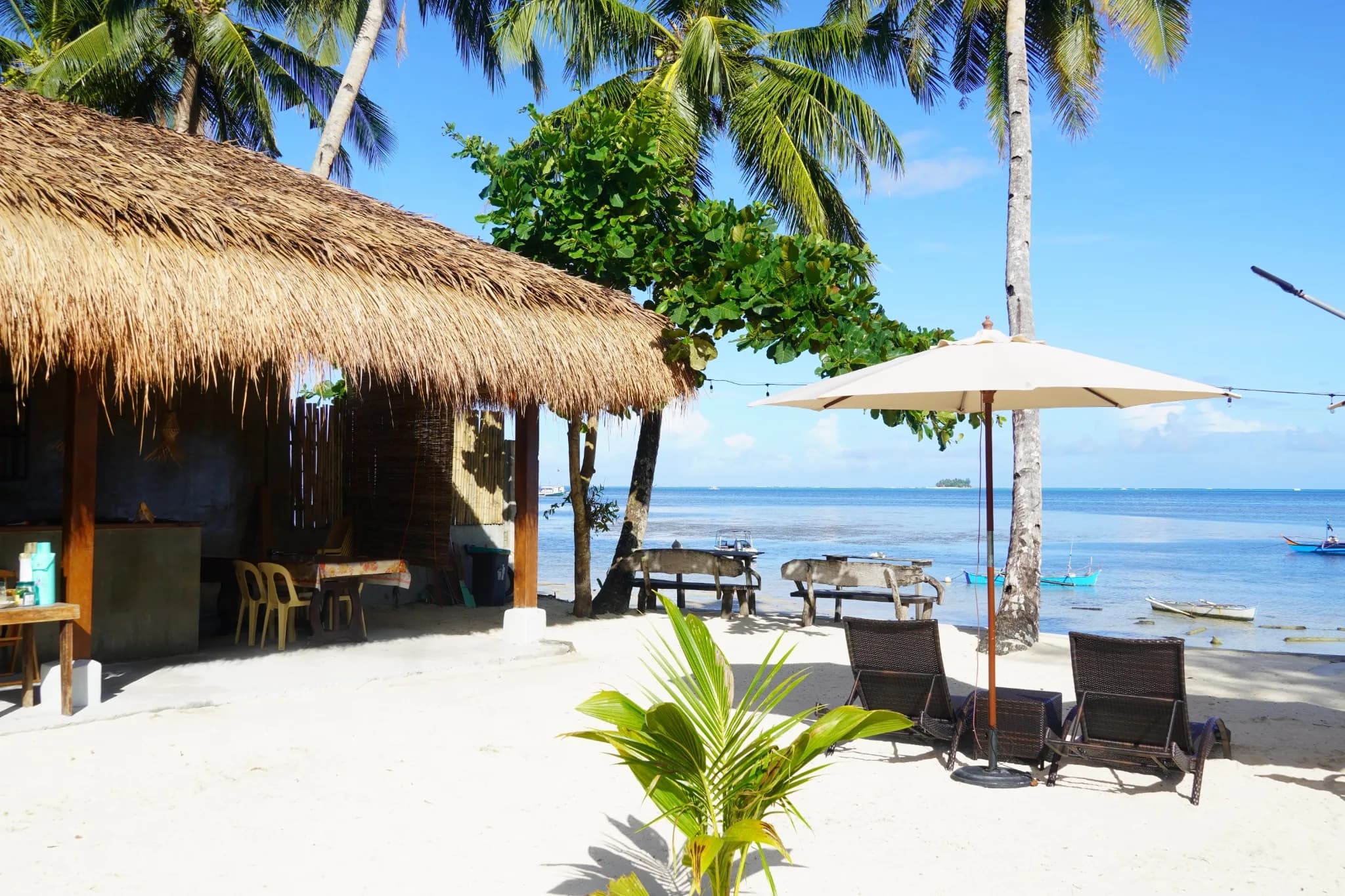 Palm-shaded sandy shore with a thatched-roof shelter, lounge chairs and boats on the sea at Aggehanan.