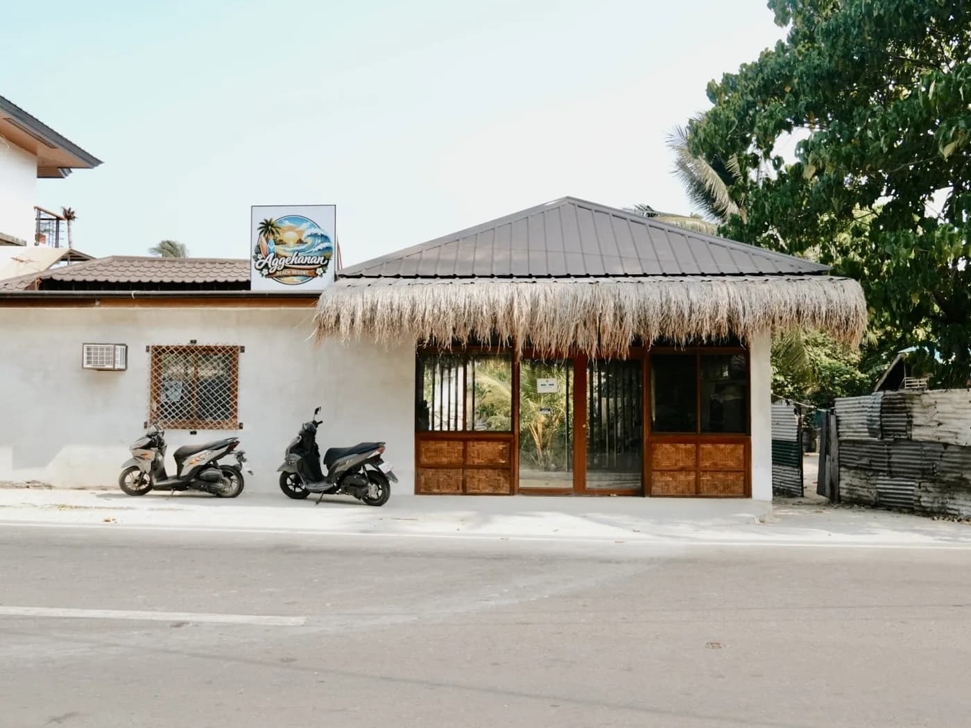 Aggehanan Beach Resort sign above the roadside entrance, with wooden doors and two parked scooters.