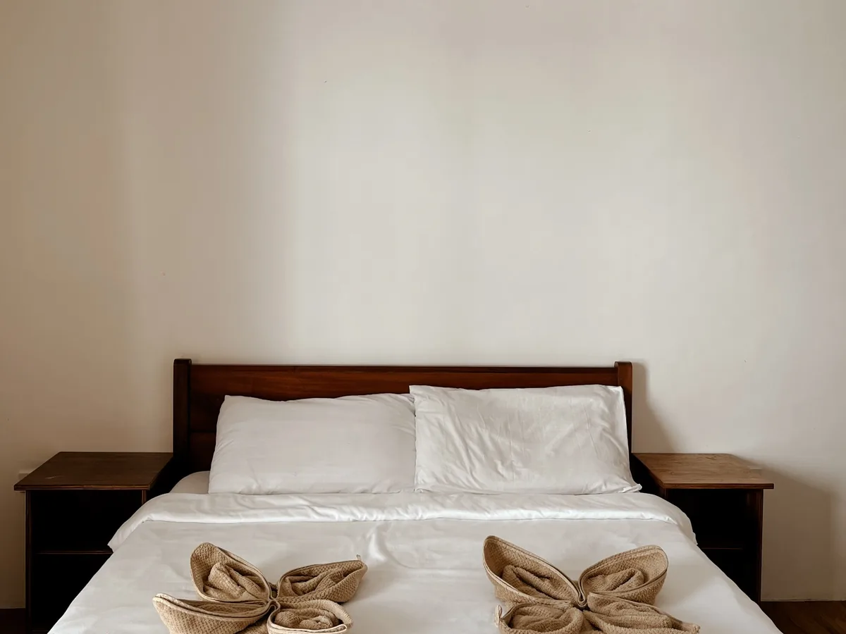 Standard Room bed with white linens, folded towels and wooden bedside tables.